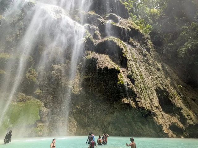 People swimming and relaxing at the base of a spectacular, towering waterfall.