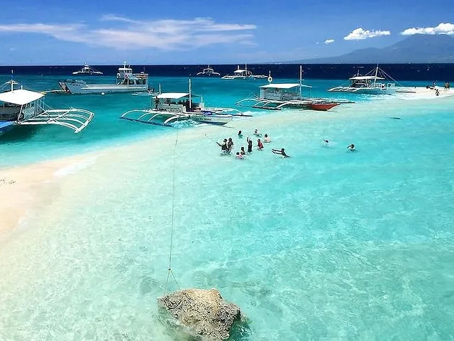 People swimming in crystal clear turquoise water near outrigger boats and a sandbar.