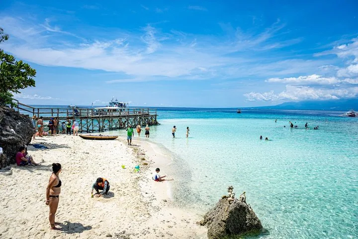 Tourists enjoying the shallow, clear waters and sandy beach near a wooden pier.